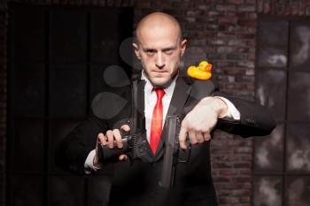 Angry contract murderer in suit and red tie aims a pistol on little toy duck. Professional secret agent concept
