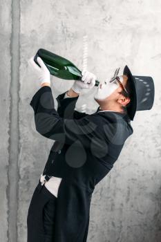 Mime theater actor drinking from a big bottle and performing a drunk man. Comedy pantomime artist in suit, gloves, glasses, make-up mask and hat