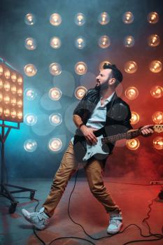 Male solo guitarist with electro guitar on the stage with the decorations of lights. Music entertainment. Bearded musican song performing 