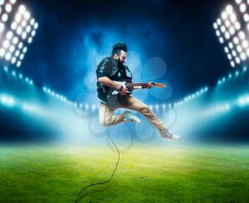 Male performer with electro guitar in a jump on the stadium stage with the decorations of lights. Music entertainment. Bearded musican song performing