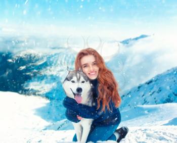 Smiling young woman hugs with husky, snowy mountains on background. Cute girl with dog. Friendship with domestic animal
