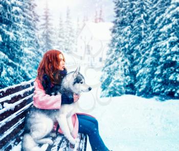 Beautiful woman sitting on the bench with siberian husky, snowy forest on background. Cute girl hugs with charming dog. Real friendship with domestic animal