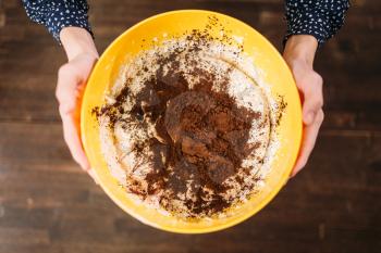 Female hands holds bowl with dough and chocolate powder against on wooden background. Sweet cake cooking preparation
