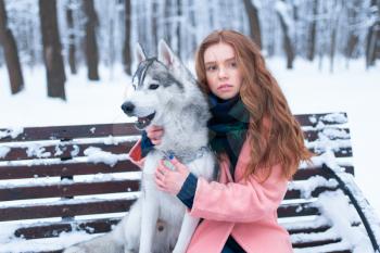 Beautiful woman sitting on the bench with siberian husky, snowy forest on background. Cute girl hugs with charming dog. Real friendship with domestic animal