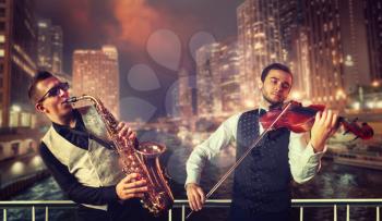 Saxophonist and violinst playing melody against night cityscape background, musical duet. Jazz-man and fiddler