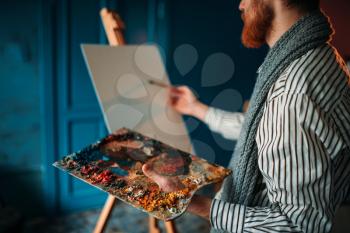 Male artist with palette and brush in hand in front of easel. Oil paint