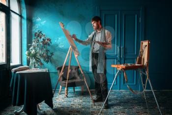 Male artist with palette and brush in hand paint on easel in front of window. Oil paint, paintbrush drawing