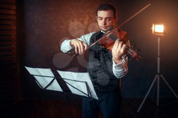 Portrait of male violinist with violin against music stand. Fiddler man with musical instrument playing in studio, solo concert training