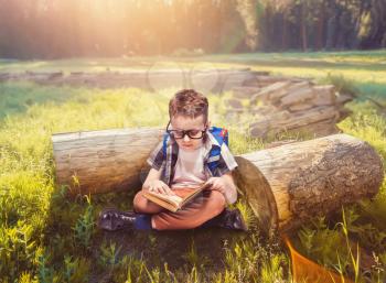 Little boy with school bag reading a book in yoga pose, green nature on background. Young pupil in glasses with backpack and textbook. Child with schoolbag sitting on a green meadow