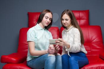 Two young women sitting on red leather couch and looking at pictures on phone
