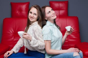 Two girls with cups of coffee sitting together on red leather couch. Women gossip