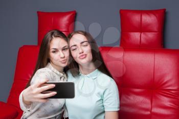 Two attractive girlfriends sitting on red leather couch and makes selfie on phone camera