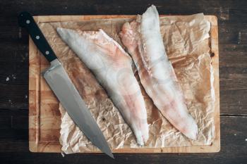 Raw fish slices and knife on cutting board covered with parchment paper, top view