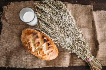 Fresh bread bun with crispy crust, wheat bunch and glass of milk on burlap background, top view