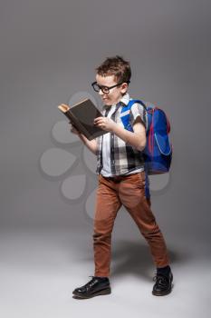 Little child with school bag reading a book, studio photo shoot. Young pupil in glasses with backpack and textbook. Boy with schoolbag