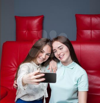 Two attractive girlfriends sitting on red leather couch and makes selfie on phone camera