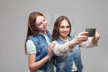 Two smiling girlfriends in denum jackets makes selfie on phone camera in studio. Female friendship. Leisure of happy girls
