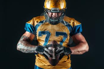 Muscular american football player in uniform and helmet holds ball in hands, black background. Contact sport