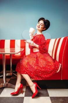 Pretty pin up girl with make-up drinks coffee in retro cafe, 50 american fashion. Red dress with polka dots, vintage style