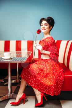 Sexy pin up woman with make-up holds red rose, dress with polka dots, vintage style. Retro cafe interior with checkerboard floor