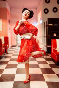 Attractive pin up girl with bright make-up sends air kiss, vintage cafe interior on background, popular american fashion 50s and 60s. Red dress with polka dot