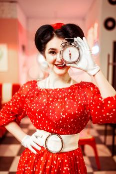 Smiling pin up girl posing with alarm clock, vintage cafe interior on background, popular american fashion 50s and 60s. Red dress with polka dots, bright make-up