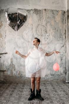 Naked freak man wrapped in packaging film holds air balloons in his hands, grunge room interior. Crazy male person in abandoned house