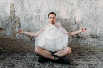 Funny freak man wrapped in packaging film sitting in yoga pose, grunge room interior. Mad male person in abandoned house, crazy guy