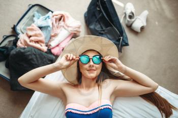Young woman lies in swimsuit and sunglasses, top view, opened suitcase with clothes on background. Fees on journey concept. Preparation for summer vacation