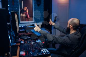 Female singer in recording room and sound engineer and at remote control panel in studio. Musician in headphones listens composition, professional music mixing