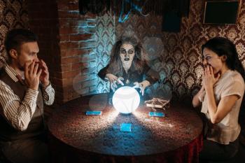 Witch reads scary magic spell over a crystal ball, young people in horror on spiritual seance. Female foreteller calls the spirits