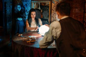 Young couple at the table with crystal ball on spiritual seance, scary witch reads the spell on background. Female foreteller calls the spirits