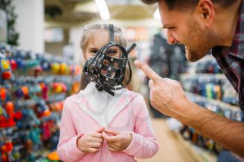 Family chooses muzzle for domestic animal in pet shop, father and little daughter having fun in petshop