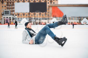 Young smiling man in skates sitting on ice, skating rink. Winter ice-skating on open air, active leisure