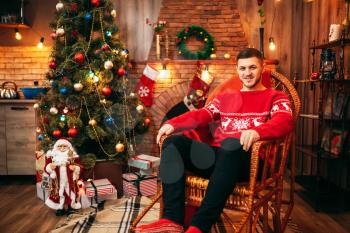 Man sitting on a chair near fireplace, christmas tree with decoration on background, xmas holiday celebration 