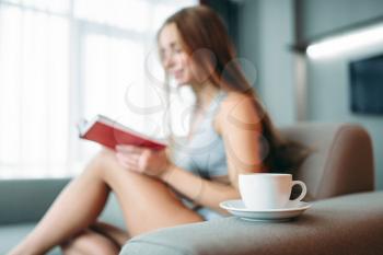 Beautiful woman in underwear sitting on the couch and reads the book against cup of coffee. Girl wake up, morning leisure