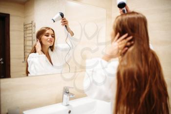 Beautiful woman in bathrobe drying hair with dryer against mirror in bathroom, morning hygiene, beauty care