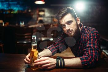 Sad man sitting at the bar counter and holds the bottle with alcohol beverage in hands. Male person in pub, alcoholism, alcohol addiction, drunkenness