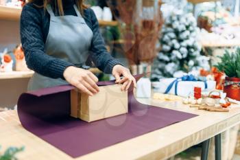 Female seller holds unwrapped carton gift box. Present wrapping papaer on the table, decoration process