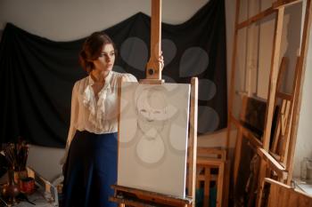Cute female artist drawing in studio. Creative paint, pencil sketch on easel, workshop interior on background