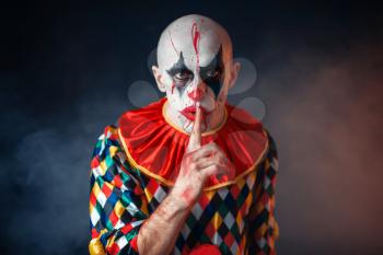 Portrait of mad bloody clown shows the quiet sign, face in blood. Man with makeup in halloween costume, mad maniac