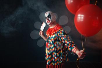 Ugly bloody clown with meat cleaver holds air balloon, horror. Man with makeup in carnival costume, mad maniac