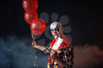 Spooky bloody clown with knife holds air balloons, horror. Man with makeup in carnival costume, crazy maniac