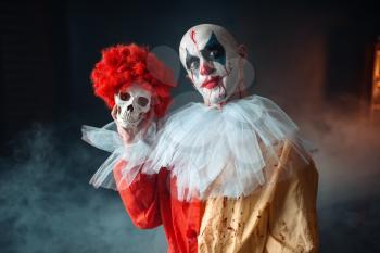 Crazy bloody clown holds human skull in red wig, horror. Man with makeup in carnival costume, mad maniac