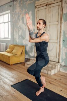 Male yoga with tattoo on hand doing balance exercise on mat in gym with grunge interior. Fit workout indoors