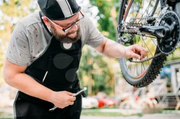Professional bicycle mechanic in apron adjusts bike chain. Cycle workshop outdoor. Bicycling sport, bearded service man work with wheel