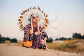 American Indian girl in native costume sitting in yoga pose, ritualistic ceremony. Headdress made of feathers of wild birds. Cherokee, Navajo culture, ethnic peoples traditions