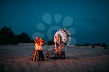 Young American Indian woman against fire, Cherokee, Navajo. Headdress made of feathers of wild birds. Night ritual