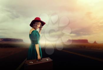 Young woman with suitcase hitchhiking, vintage style fashion. Female traveler in retro hat and dress on roadside. Road adventure