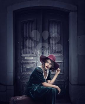Young woman sits on suitcase against grunge wooden door, vintage style fashion. Female traveler in retro hat and dress. Summer journey 